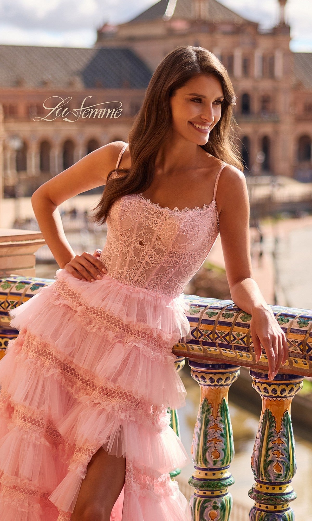 A woman wearing the Long Prom Dress: La Femme 33493, featuring a pink lace corset bodice and tiered ruffle skirt, stands by a colorful ornate railing and smiles with historic architecture in the background.